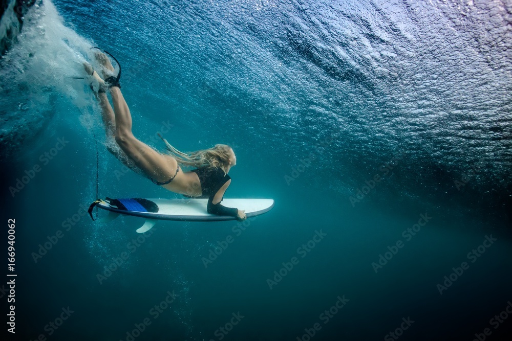 Blonde girl Surfer holding white surf board Diving Duckdive under Big Beautiful Ocean Wave. Turbulent tube with air bubbles and tracks after sea wave crashing. - obrazy, fototapety, plakaty 