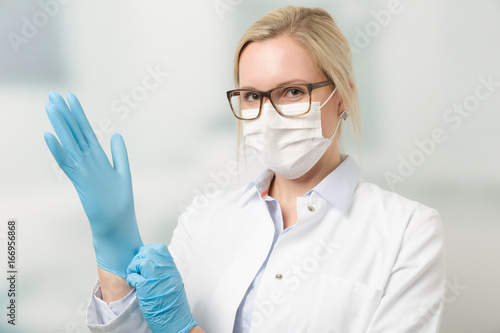 Fotografie, Obraz Junge hübsche Ärztin mit Mundschutz zieht Handschuhe an