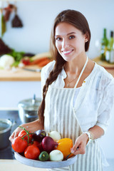 Wall Mural - Smiling young woman holding vegetables standing in kitchen