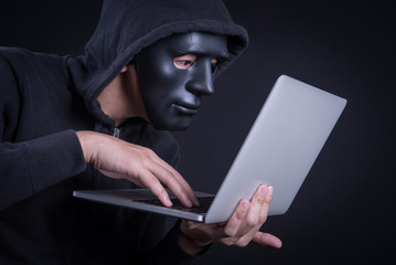 Wall Mural - Anonymous male hacker using black mask to cover his face carrying laptop computer. Internet security and cyber attack concepts.