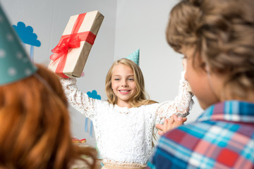 adorable happy girl holding birthday gift and looking at friends standing on foreground