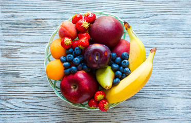 Bowl of fresh fruit with banana, apple, strawberries, apricots, blueberries, plums, whole grains, forks, top view