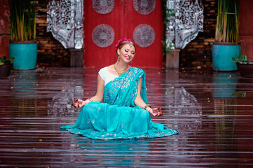 Wall Mural - Beautiful young caucasian woman in traditional indian clothing with bridal makeup and jewelry. Bollywood dancer in Sari and henna on hands meditating at temple garden.