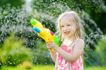 Wall Mural - Adorable little girl playing with water gun on hot summer day. Cute child having fun with water outdoors.