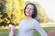 Sporty mature woman hold bottle with water outdoor on sunny day in the park