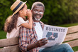 african american girl whispering to her grandfather while he reading business newspaper on bench