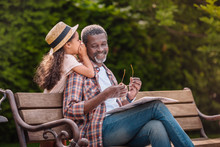 Little Adorable African American Grandchild Whispering To Her Smiling Grandfather While Sitting On Bench In Park