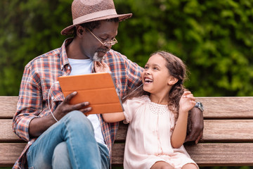 happy african american grandchild and her grandfather listening music on digital tablet while sitting on bench in park