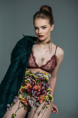 Wall Mural - young sensual stylish model posing in floral skirt, lace bra and green fur coat for fashion shoot on grey