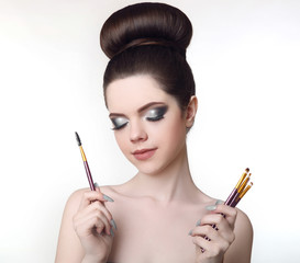 Makeup artist. Pretty teen girl with cute bun hairstyle and fashion beauty makeup, brunette holding brushes in hand isolated on white studio background.