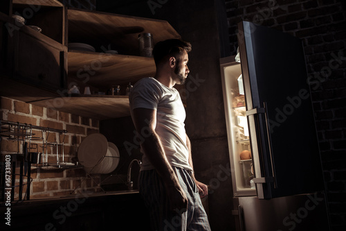 Fotografie, Obraz side view of bearded young man in pajamas looking at open refrigerator at night