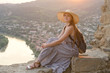 Beautiful girl with backpack in a wide hat sitting on background of the river, mountains and the city below