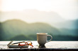  A coffee cup, eyeglasses and a note book on wooden table with sunrise and mountain  bokeh background. A start of new day with hot beverage. Business concept.