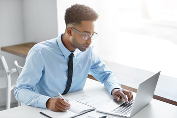 Handsome young dark-skinned manager with stubble wearing eyeglasses and blue shirt with tie having concentrated look while working on presentation using laptop computer, writing down in notebook