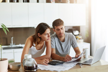 European couple sitting together indoors over home interior, calculating finances at home, looking seriously in documents. Young female with her husband doing home budget while sitting in kitchen