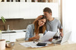 Frustrated woman studying sheet of paper in kitchen, drinking coffee and trying to find way to pay off all family debts; her husband kissing her on head, saying: Everything is going to be alright