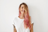 Fototapeta  - Horizontal portrait of pleasant-looking Caucasian female with long hair, pink on tips, having tattooes on arms, wearing white casual T-shirt, covering her face with hair, looking happily in camera
