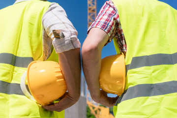 Poster - Close-up rear view of the hands of two workers wearing reflective safety vests while holding yellow hard hats outdoors on the construction site