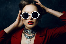 Indoor Studio Close Up Portrait Of Young Beautiful Woman Wearing Stylish Round White Sunglasses, Big Silver Boho Necklace, Red Jacket. Model With Red Lips, Long Hair. Female Beauty, Fashion Concept