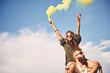 © Connect Images - Young boho woman holding yellow smoke flare on boyfriend's shoulders at festival