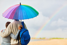 Back View Of Tourists With Backpack Standing Under Colorful Umbrella In Rural Environment