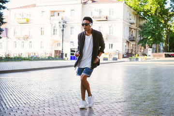 Portrait of young stylish hipster black man in white headphones and sun glasses dancing outdoor in city centre, having some fun