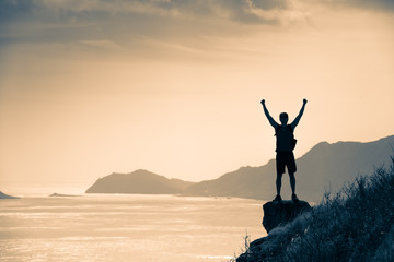 victory, winning, adventure, goals concept. man standing on mountain with arms in the air celebratin