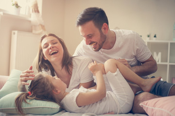 Wall Mural - Parents having fun with their little daughter on bed. Family spending time at the morning.