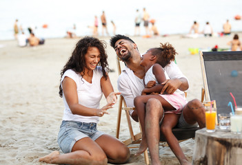 Wall Mural - Young mixed race family sitting and relaxing  at the beach on beautiful summer day.Daughter lies in father lap and laughing.