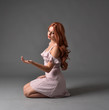 full length portrait of a pretty girl with long copper hair, wearing a simple purple dress. seated pose against a grey background.