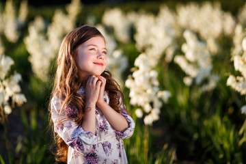 Wall Mural - A portrait of a cute little girlwith long hair in outside at sunset in the field of white yucca flowers having fun with raised hands up