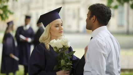 Wall Mural - Female graduate in academic regalia with flowers talking to boyfriend, education