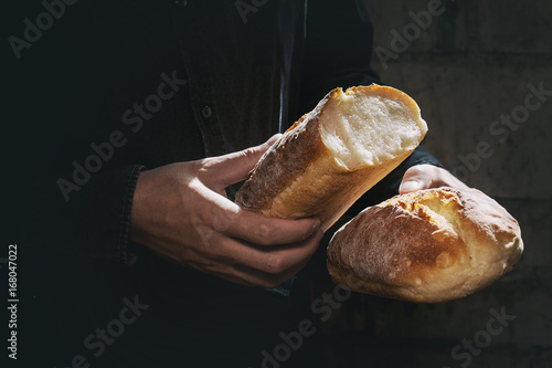 Plakat Bochenek świeży piec chlebowy chleb w mężczyzna rękach w świetle słonecznym. Rustykalne światło dnia w ciemnym pokoju. Stonowany obraz