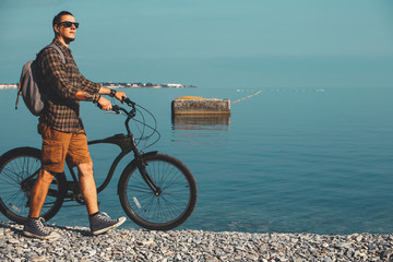 Poster - Young Man Cyclist In Sunglasses With Bicycle Walking On Coast And Enjoying View Of Sea. Holiday Travel Activity Concept