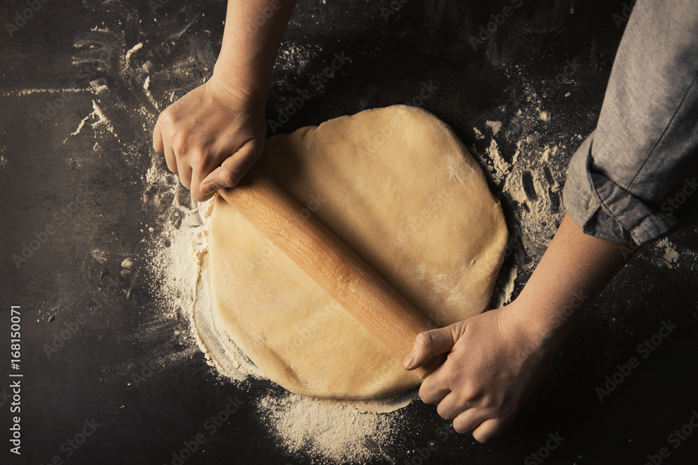 Woman rolling out raw dough on dark table