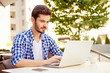 © deagreez - Stylish freelancer is working on his computer, sitting outdoors on a summer day, with cup of coffee, nice city view and green tree behind him