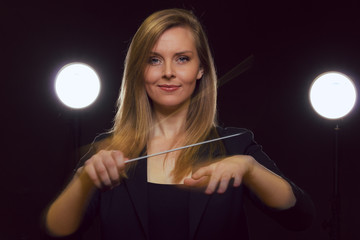 Wall Mural - Young caucasian woman conductor poses with baton in a dark space with stage lights