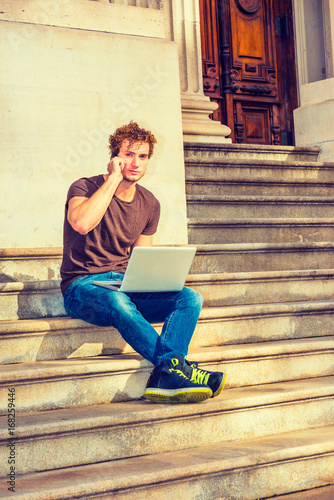 Fotografie, Obraz Young Man working on laptop computer, talking on cell phone, sitting on stairs i
