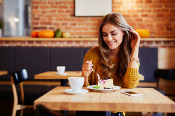 Gorgeous smiling young woman eating cake and drinking coffee at a cafeteria