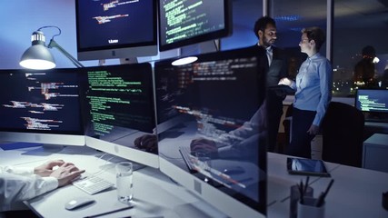 Experienced male cyber security professional working on multi-screen computer in office at night and talking on mobile phone while businesswoman and businessman discussing something on tablet