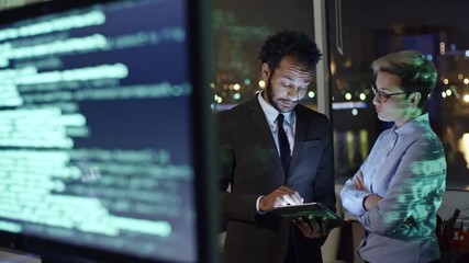 Tracking shot of computer monitors with programming codes and two colleagues discussing something on digital tablet while working in office at night