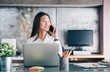 © weedezign - Asian businesswoman talking on mobile phone in front of laptop computer and looking out of window at office,Office lifestyle concept.