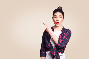 Wall Mural - Young surprised girl with casual style and bun hair pointing her finger sideways, demonstrating something on beige blank wall with copy space for your information or promotional content.