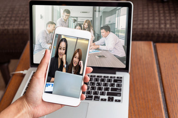 Hand holding the technology smart mobile phone having video conference via monitor display for working in the Loft cafe workplace, entrepreneur business and technology concept