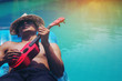 The young man relaxes and plays Ukulele on a rubber float floating in an outdoor pool in the sun.