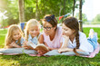 © pressmaster - Teacher and three little girls reading stories from book while relaxing on grass