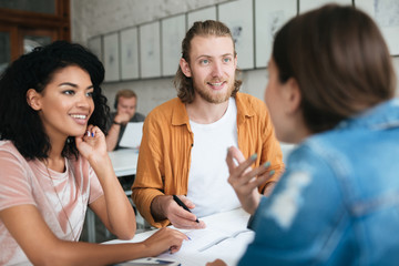 Wall Mural - Young man and two girls working together in office. Group of students studying together in classroom. Portrait of cool young people happily talking and discussion something together