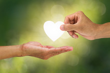 Hand holding and giving white heart to receiving hand on blurred green bokeh background, helping hand concept