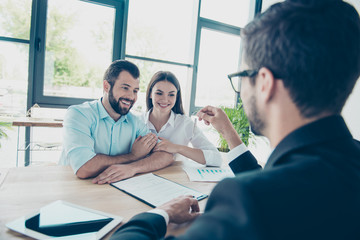 Happy young couple is embracing, getting a key from their future apartment from a broker, and signing contract, all are dressed in formal outfits, nice light office design