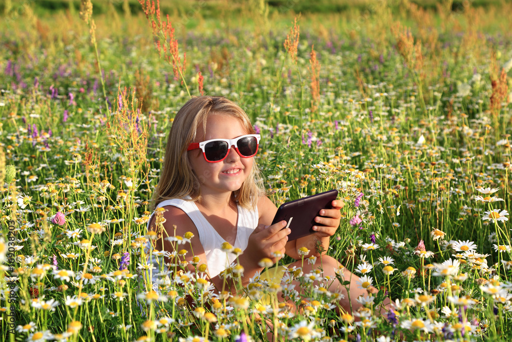 Śliczna uśmiechnięta dziewczynka bawi się tabletem wśród polnych kwiatów. - obrazy, fototapety, plakaty 